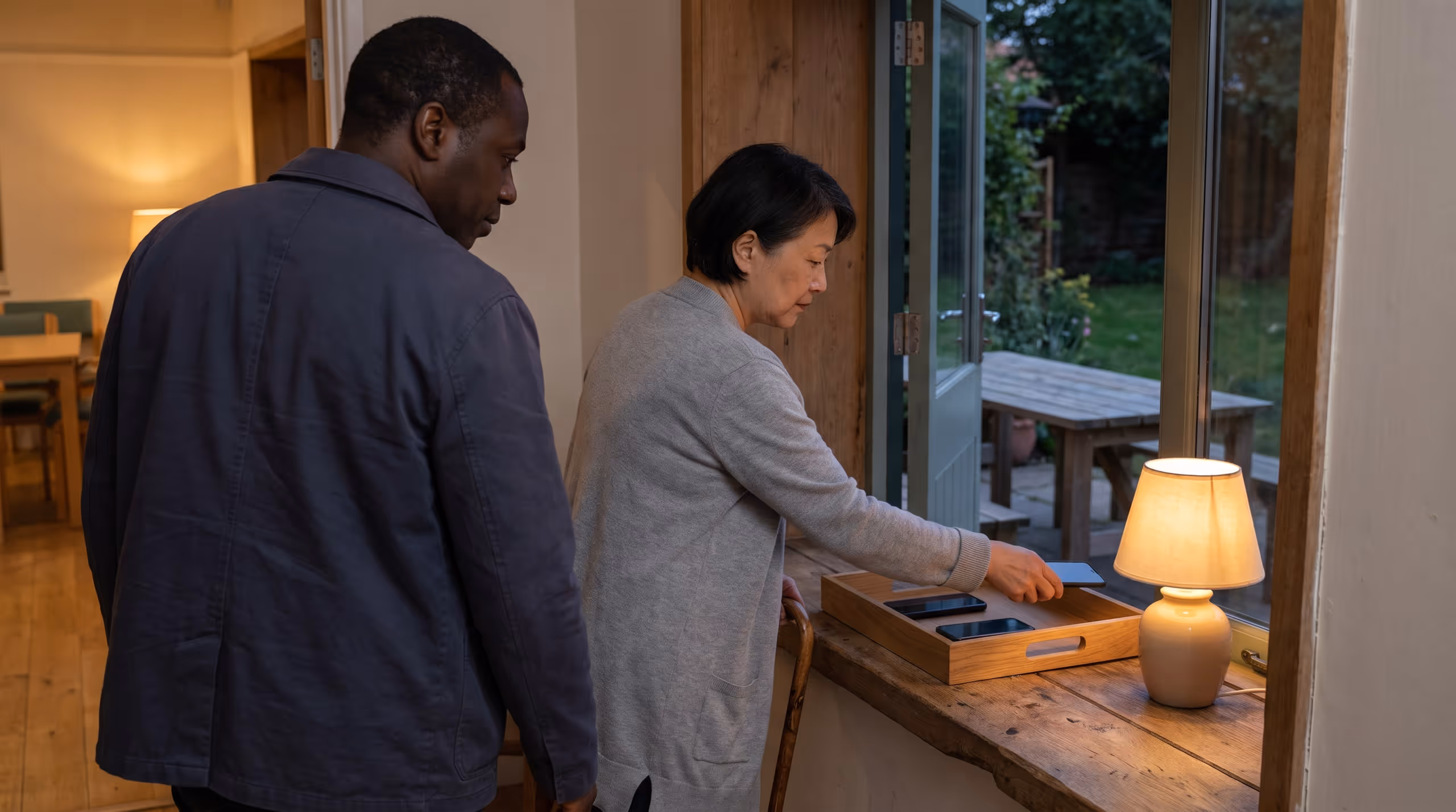 Two adults voluntarily set phones in a tray beside a doorway opening onto a garden.